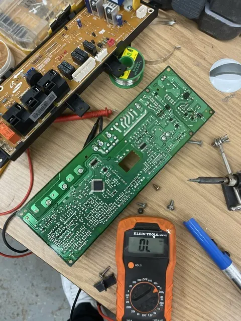 Two appliance control boards on a workbench with a multimeter